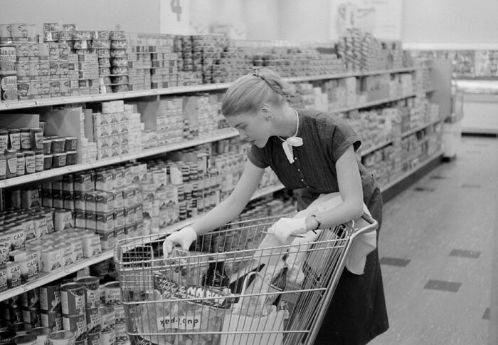 1940s housewife wearing gloves, shopping for canned goods in a grocery store aisle with a metal cart.