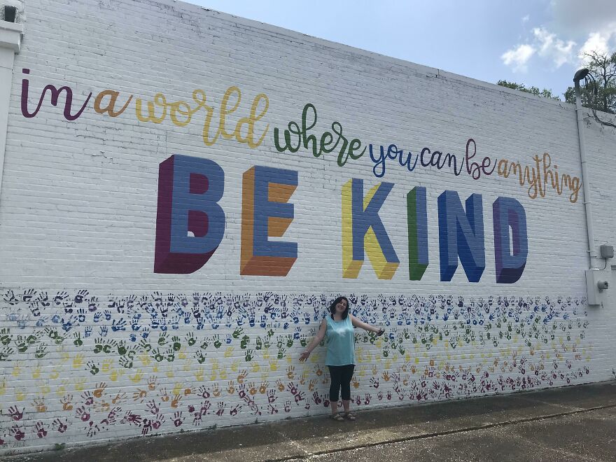 Colorful mural with perfect handwriting example saying be kind on white brick wall with person standing below it outdoors