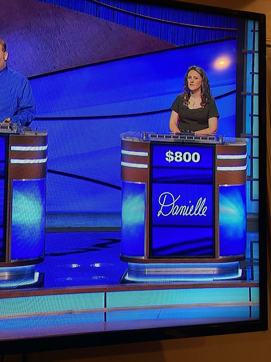 Contestant standing at a game show podium with perfect handwriting displaying the name Danielle in elegant script.