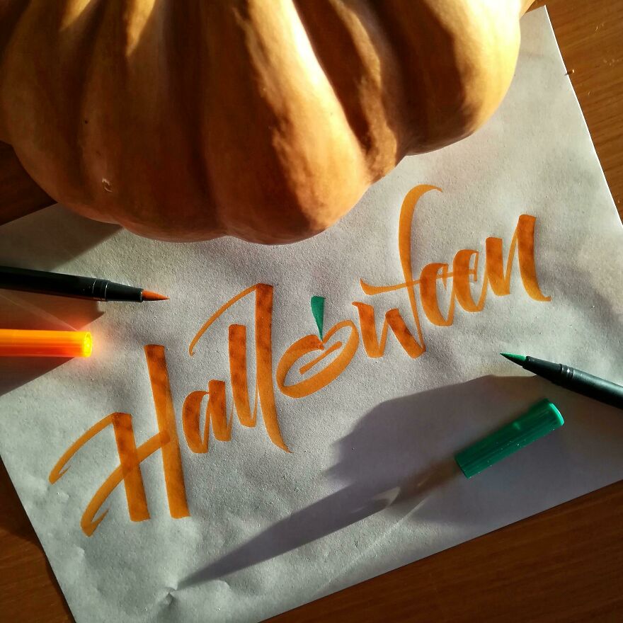 Handwriting example of the word Halloween in orange and green brush lettering with a pumpkin and markers on a wooden table.