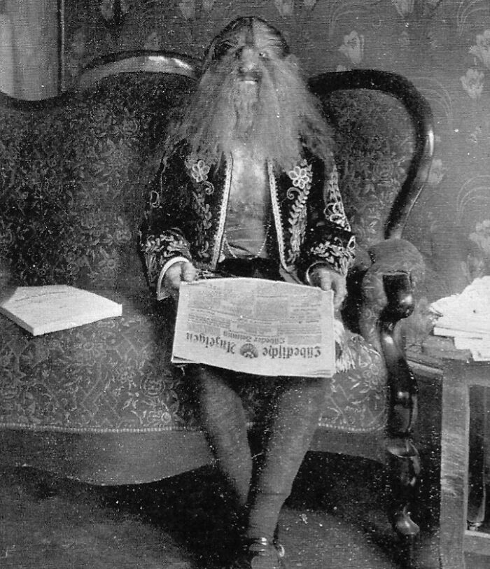 Black and white photo of a person with a grotesque mask sitting on a Victorian couch holding a newspaper, creepy vintage photo.
