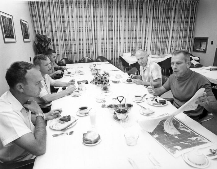 Apollo mission astronauts seated at a dining table, sharing a meal and discussing maps in a black and white photo.