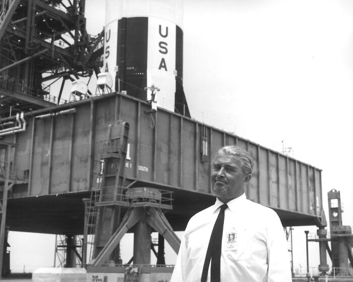 Man in white shirt and black tie standing near Apollo mission rocket launch platform with USA markings.
