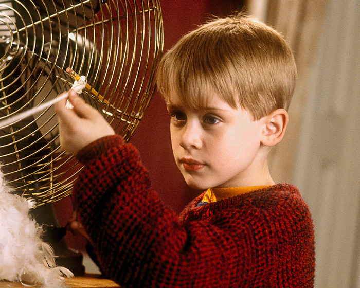 Young beloved 90s child star focused, wearing red sweater, interacting with a metal object in an indoor setting.