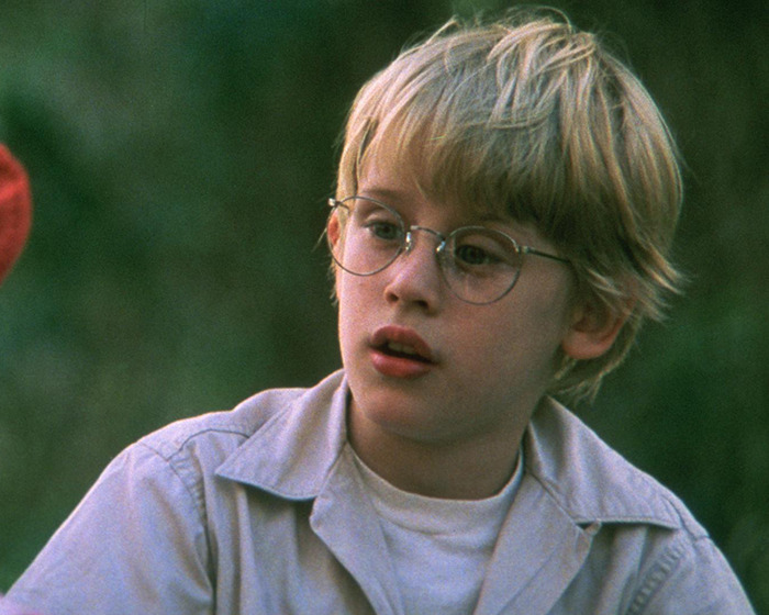 90s child star wearing round glasses and light shirt, looking thoughtful outdoors with green blurred background 90s child star wearing round glasses and light shirt, looking thoughtful outdoors with green blurred background