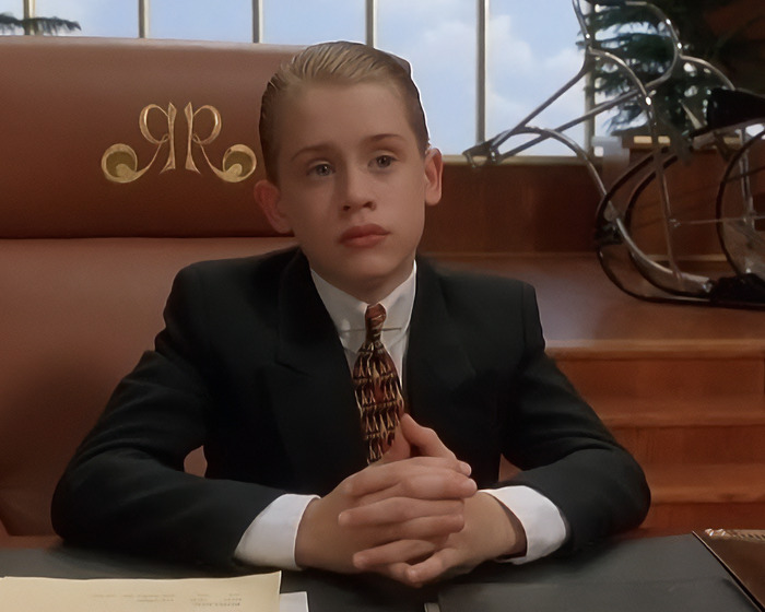 90s child star wearing a suit and patterned tie, sitting in a leather chair with a serious expression indoors. 90s child star wearing a suit and patterned tie, sitting in a leather chair with a serious expression indoors.
