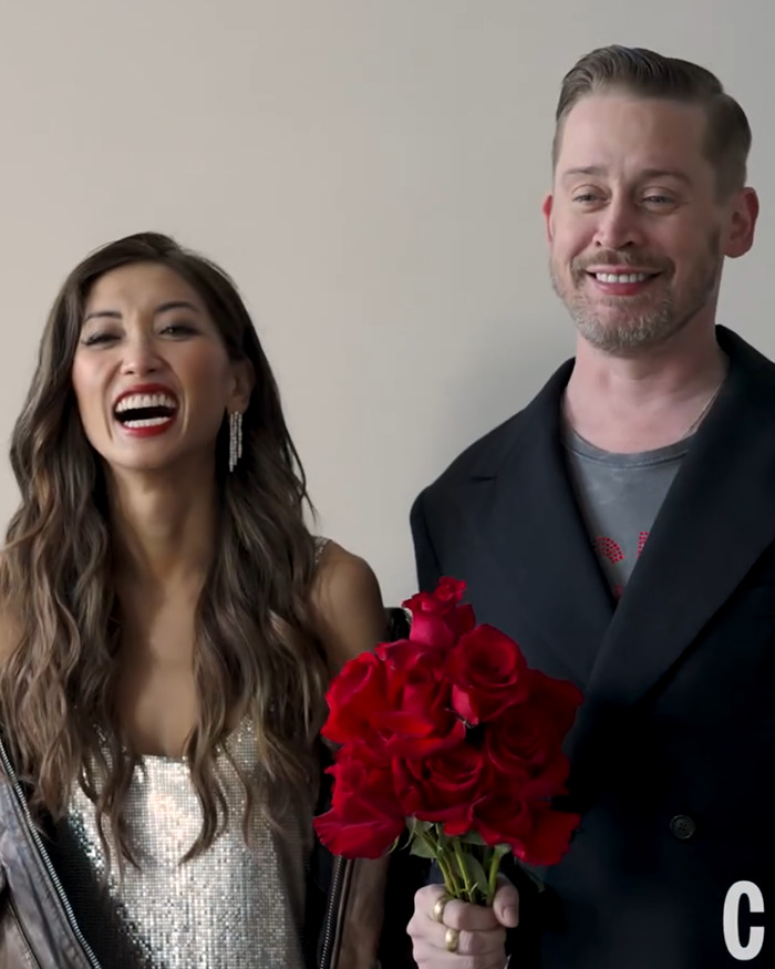 Man holding a bouquet of red roses beside a smiling woman wearing a silver dress in a casual indoor setting. Man holding a bouquet of red roses beside a smiling woman wearing a silver dress in a casual indoor setting.