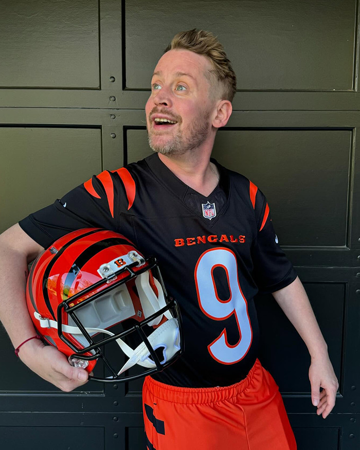 Man in Bengals jersey and shorts holding a football helmet, looking surprised in a rare outing of a beloved 90s child star.