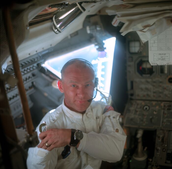 Apollo mission astronaut inside spacecraft cabin wearing headset and white suit, surrounded by control panels and equipment.