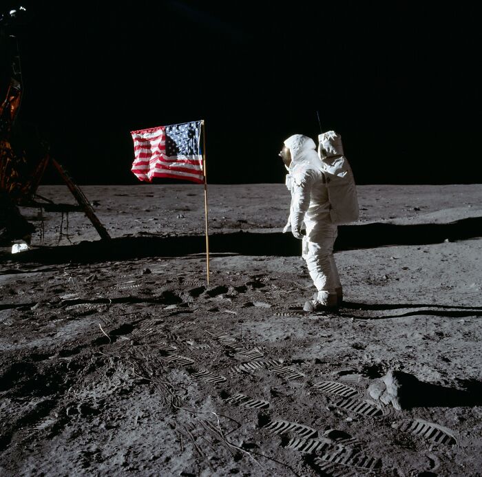 Astronaut standing on the moon next to the American flag during an Apollo mission with visible lunar footprints.
