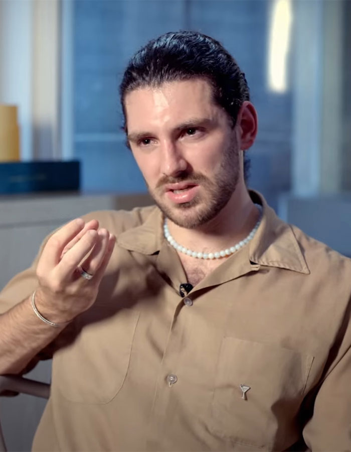 Young man in a brown shirt and pearl necklace, speaking seriously in an indoor interview about anti-vax mom controversy.