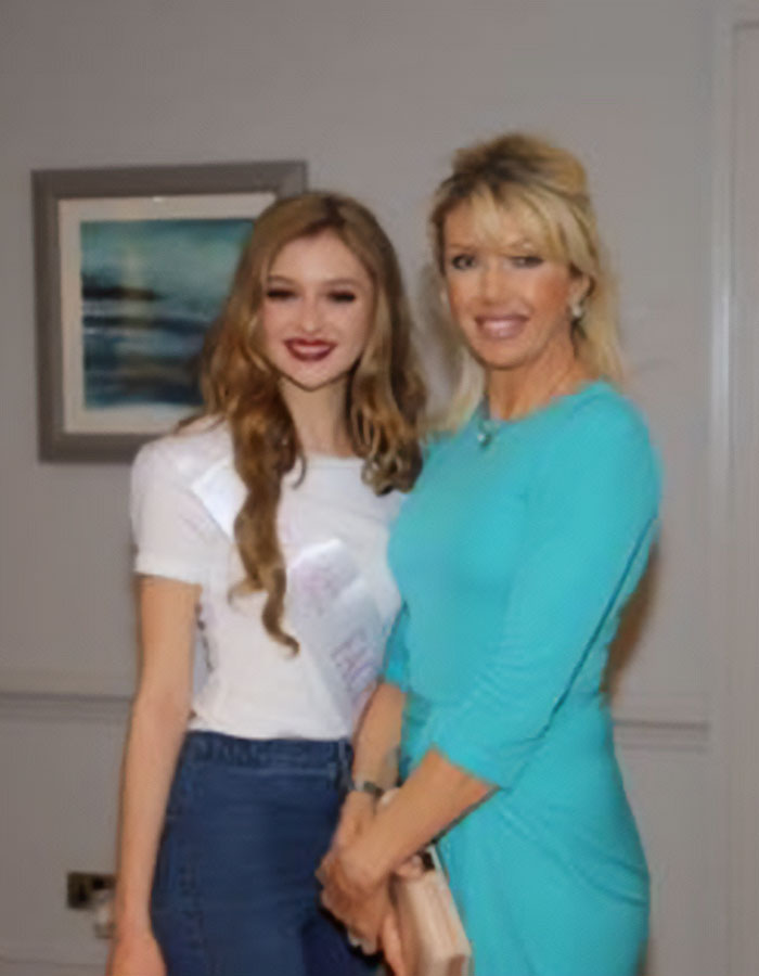 Anti-vax mom with her daughter, Cambridge 23YO graduate, standing together indoors smiling at the camera.