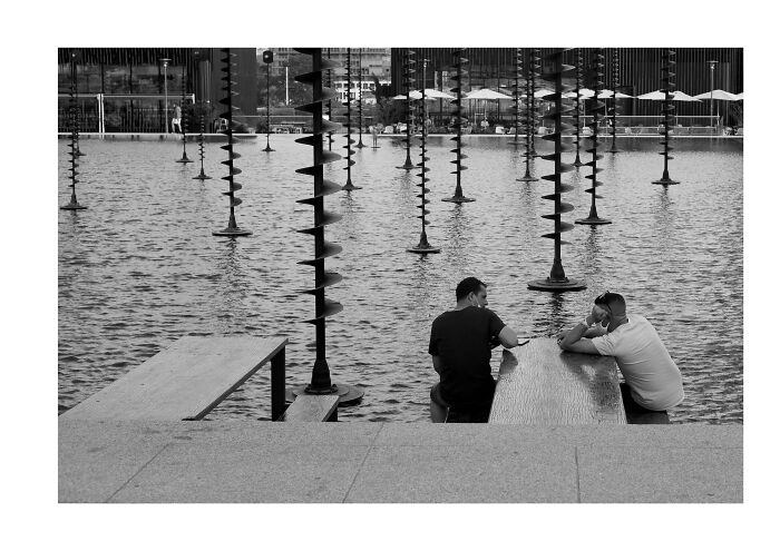 Two men sitting at a table near water with spiral sculptures in a cinematic black and white photo by Adriana Ferrarese.