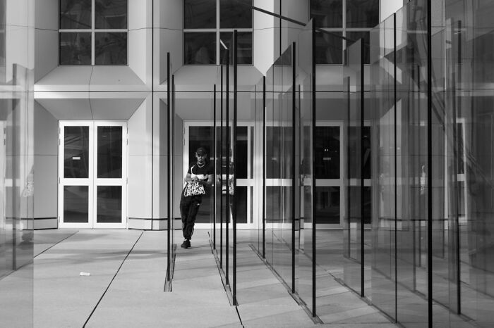 Black and white cinematic photo of a person walking between glass panels reflecting urban architecture and everyday life.