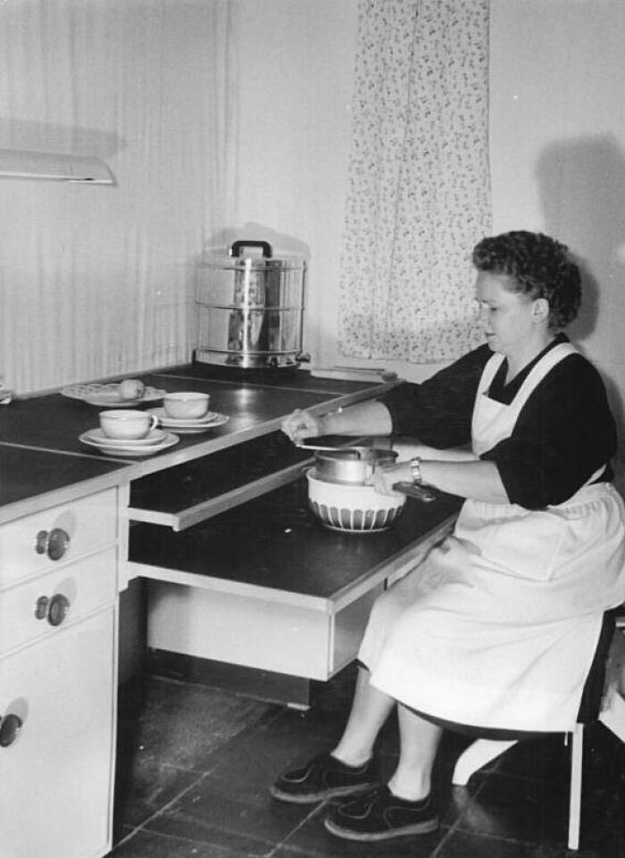 1940s housewife in apron stirring bowl at kitchen counter with tea cups and vintage appliance nearby