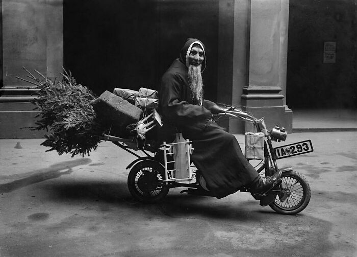 Old man with long beard riding a vintage motorbike carrying a Christmas tree and packages in a historic black and white photo.