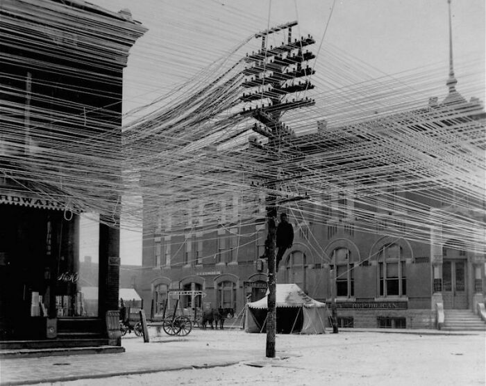 Historic photo showing a confusing network of telegraph lines on poles in a snowy town square from history.