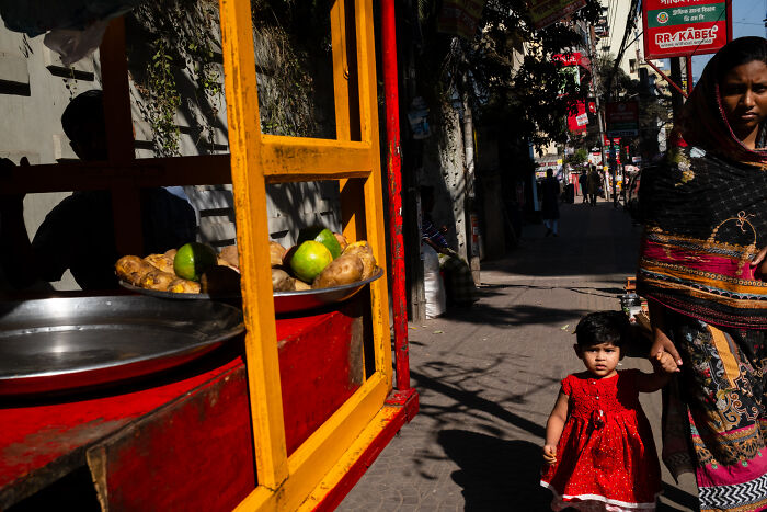 Candid street moment of a child in a red dress holding an adult’s hand on a sunlit urban sidewalk.