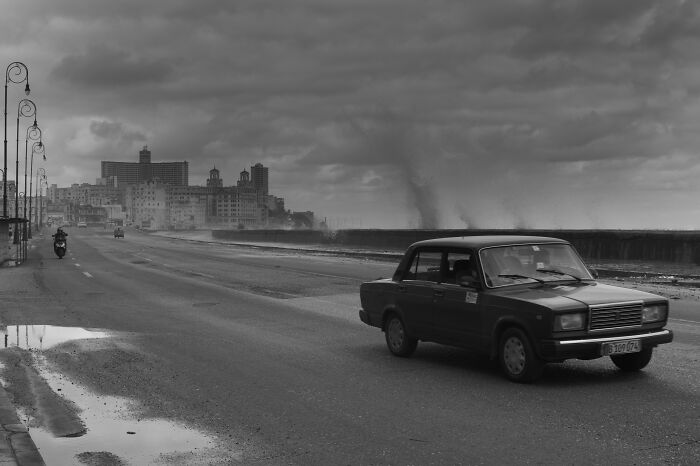 Black and white cinematic photo of an old car driving by the ocean under a moody cloudy sky.