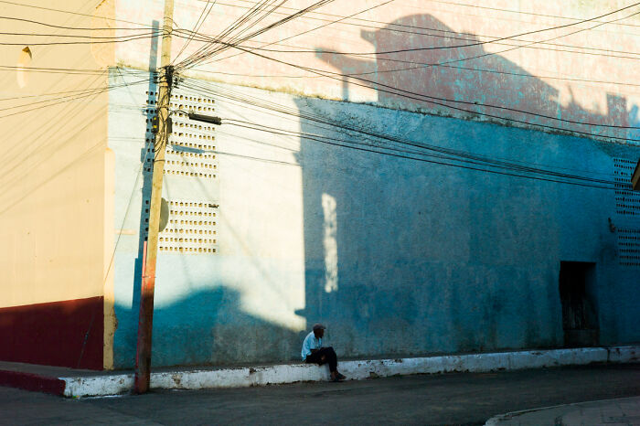 Man sitting alone on curb under dramatic shadows, captured in a cinematic photo by Adriana Ferrarese showcasing everyday life.