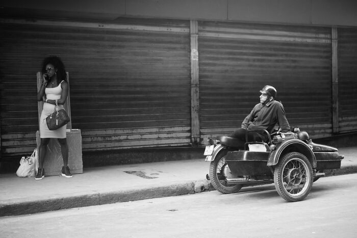 Black and white cinematic photo showing a woman standing and a man sitting on a vintage motorcycle sidecar.