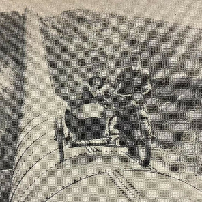 Vintage photo of a man riding a motorcycle with a sidecar on a large industrial pipe in a hilly area, historical image.
