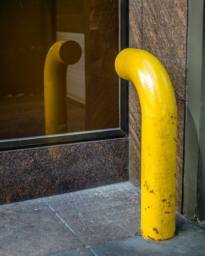 Yellow bollard beside a building reflected in a window, capturing a street photographer’s eye for magical city life coincidences.