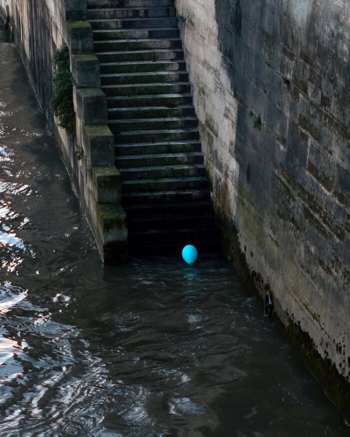 Blue balloon floating near mossy stairs by concrete wall in city river, capturing street photographer’s magical city life coincidence.
