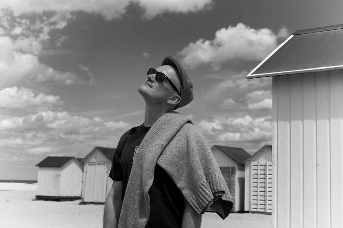 Black and white cinematic photo of a man in sunglasses and cap on a beach with cabins under a cloudy sky