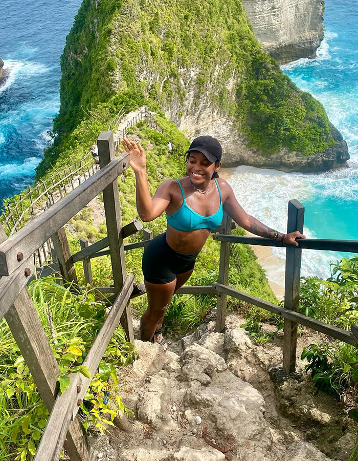 Woman hiking steep trail along cliffside near active volcano with ocean waves in background during sunny day Woman hiking steep trail along cliffside near active volcano with ocean waves in background during sunny day