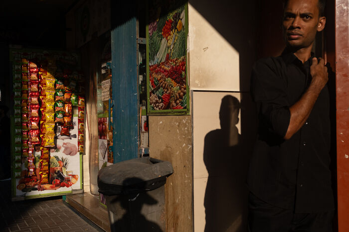 Man standing in a sunlit street corner near a shop, captured in a candid street moment full of emotion by photographer.