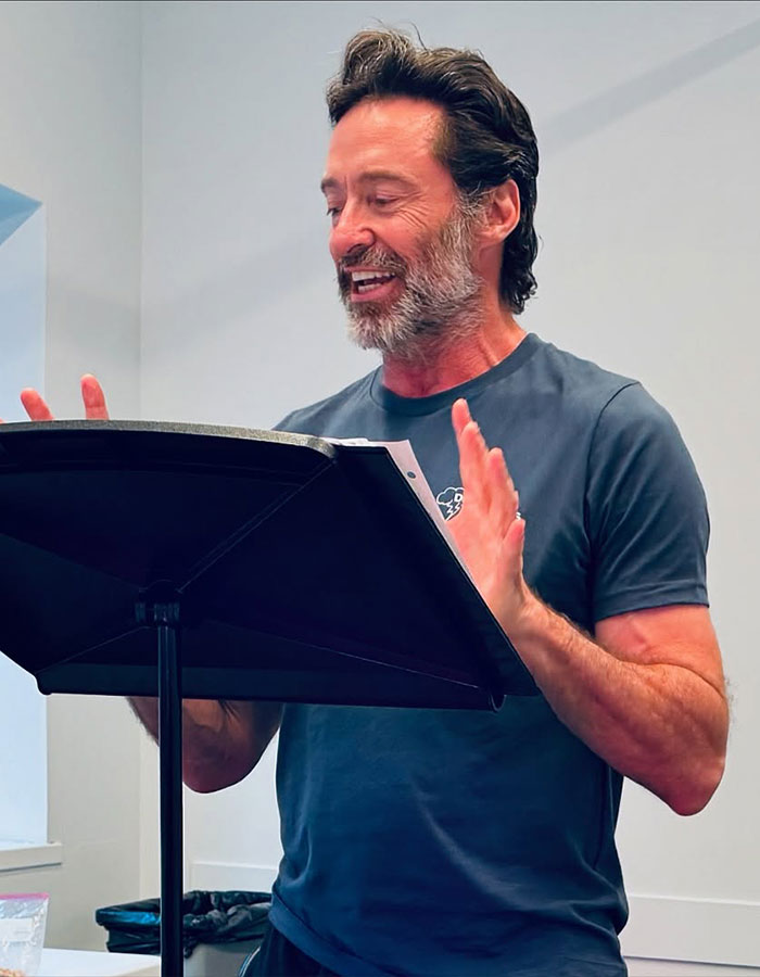 Hugh Jackman speaking animatedly at a music stand, showing expressive hand gestures and casual attire indoors. Hugh Jackman speaking animatedly at a music stand, showing expressive hand gestures and casual attire indoors.