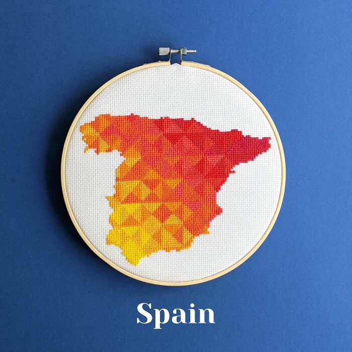 Cross-stitched map of Spain in warm gradient colors displayed in an embroidery hoop on a blue background.