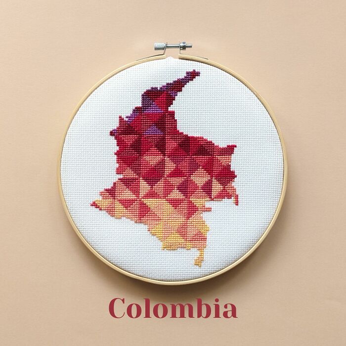 Cross-stitched map of Colombia in warm red and orange shades displayed in a wooden embroidery hoop on beige background.