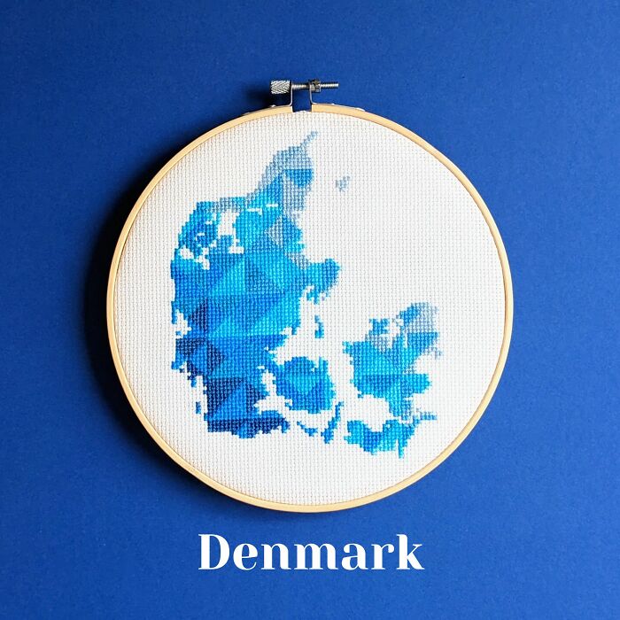 Cross-stitched map of Denmark in blue shades displayed in an embroidery hoop on a blue background.
