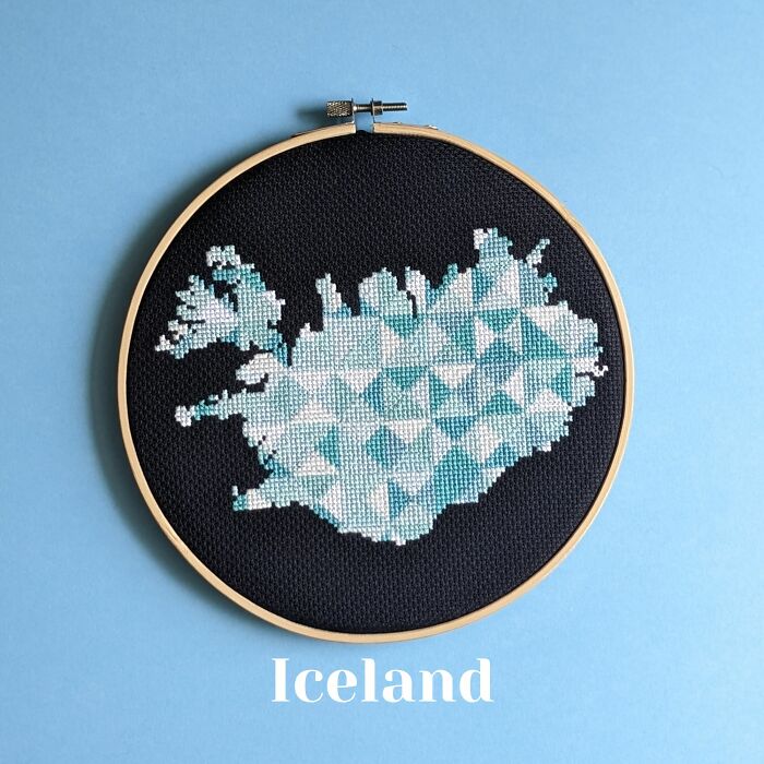 Cross-stitched map of Iceland in shades of blue and white on black fabric inside a wooden embroidery hoop.