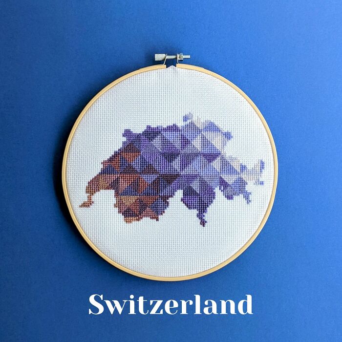 Cross-stitched map of Switzerland in shades of purple and brown displayed in an embroidery hoop on a blue background.