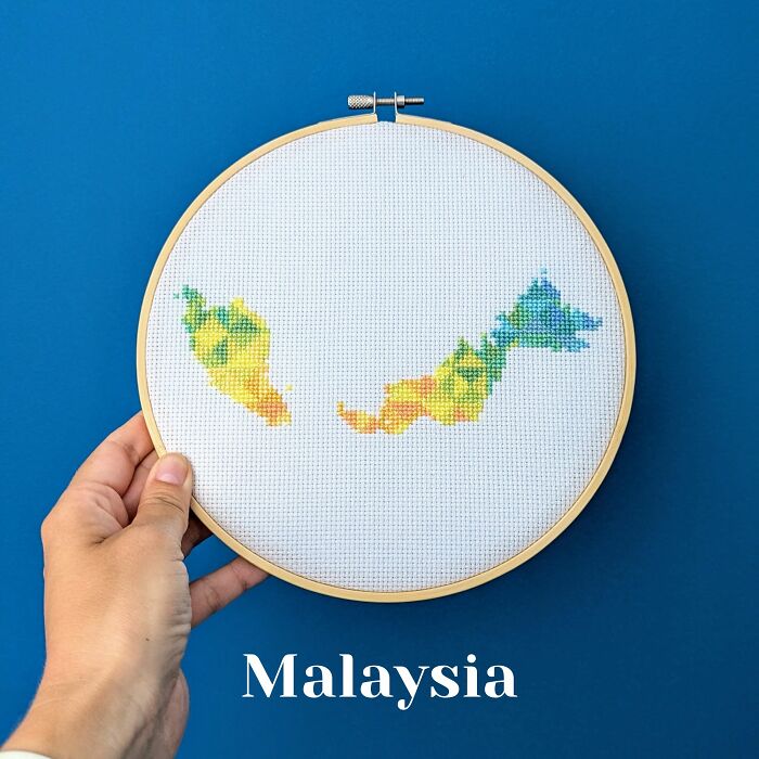 Hand holding a cross-stitched map of Malaysia in an embroidery hoop against a blue background.