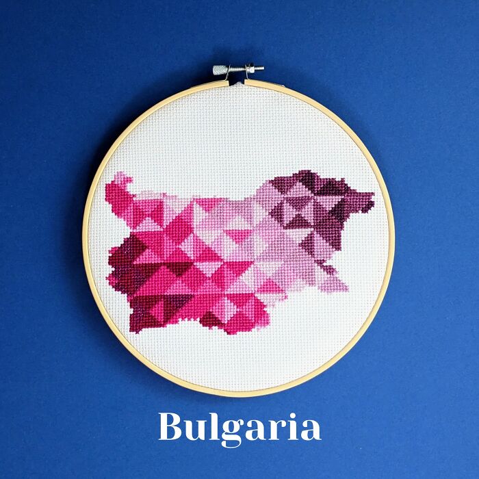 Cross-stitched map of Bulgaria in shades of pink and purple displayed in an embroidery hoop on a blue background.