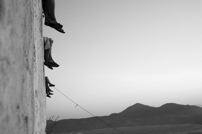 Black and white cinematic photo by Adriana Ferrarese showing people’s legs dangling over a wall with mountains in the background.