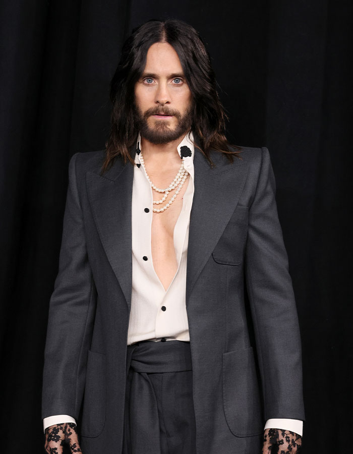 Jared Leto wearing a dark suit and pearl necklace, posing against a black backdrop amid underage claims exposé. Jared Leto wearing a dark suit and pearl necklace, posing against a black backdrop amid underage claims exposé.