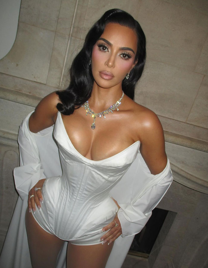 Kim Kardashian posing in a white corset and jewelry in a glamorous setting, with fans discussing her plastic surgeries. Kim Kardashian posing in a white corset and jewelry in a glamorous setting, with fans discussing her plastic surgeries.