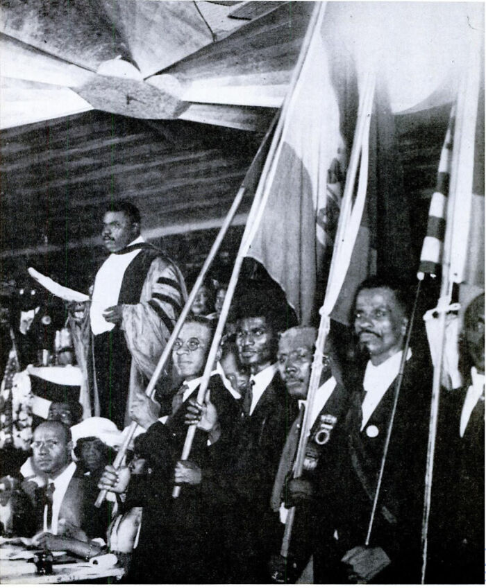 Black leaders and men holding flags at a Harlem Renaissance event with a speaker addressing the crowd indoors.