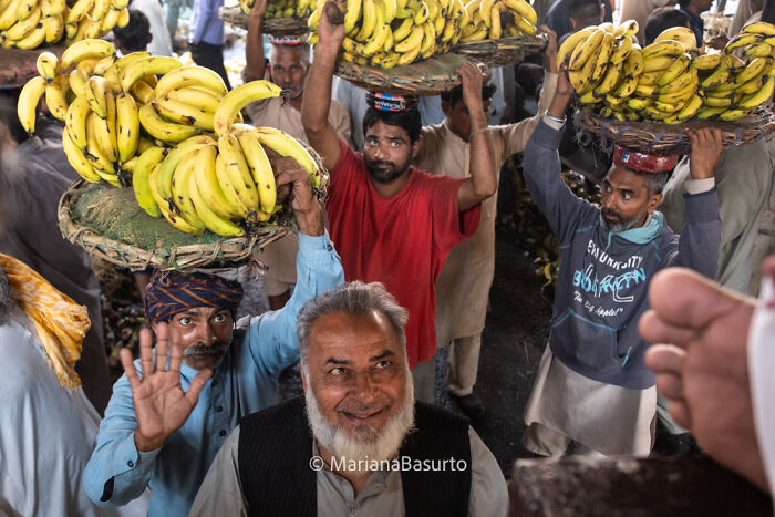 Men carrying large baskets of bananas on their heads at a busy market, showcasing unseen realities captured by a photographer.