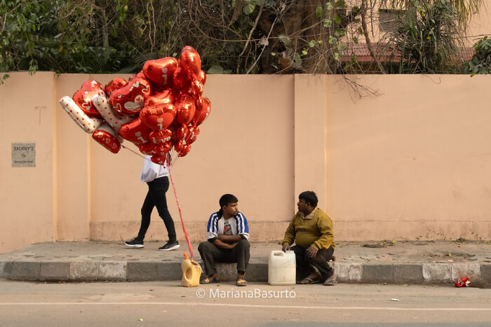 Two men sitting on a sidewalk with jerry cans, while a person walks past holding red heart-shaped balloons in a street scene.