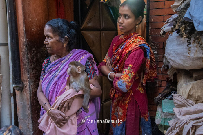 Two women in colorful traditional clothing stand in a narrow alley as one holds a monkey, revealing unseen realities.