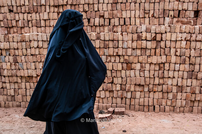 Woman in a black garment walking in front of stacked bricks, capturing unseen realities of our world through photography.