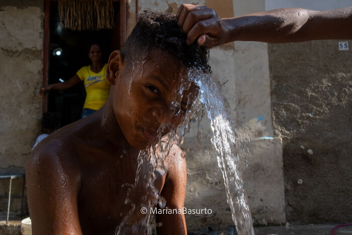 Boy washing his hair with water outside a rustic home, revealing unseen realities of our world through photography.