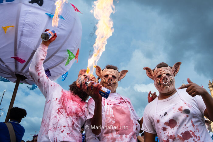 Three people in pig masks and blood-stained shirts performing fire breathing at a colorful outdoor event.
