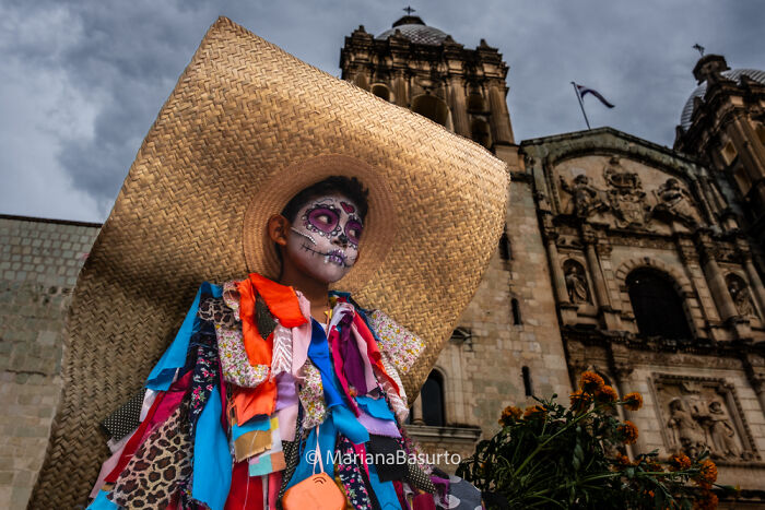 Child with painted skull face and colorful costume wearing a large straw hat in front of historic church, revealing unseen realities.